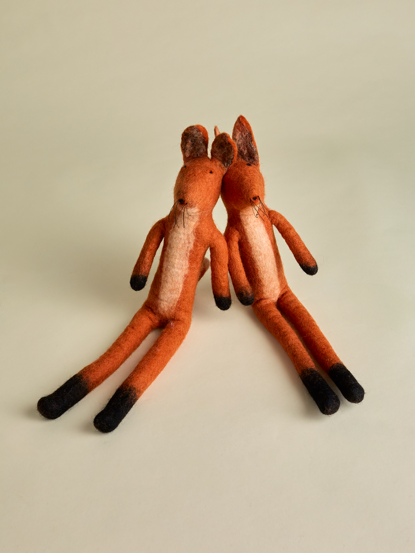 Two sitting felt foxes that are orange with dark colored feet and paws on neutral background