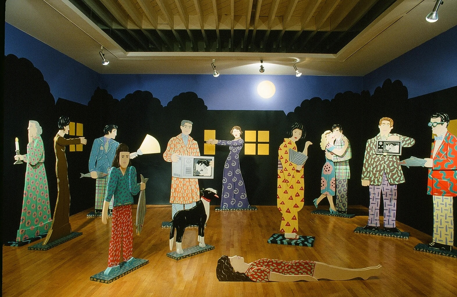A staged interior scene with multiple stylized cardboard or cutout human figures in colorful patterned outfits arranged on a wooden floor in front of a painted backdrop showing a dark night sky, trees, and windows. A black dog figure stands near the center, and a reclining figure lies on the floor in front.