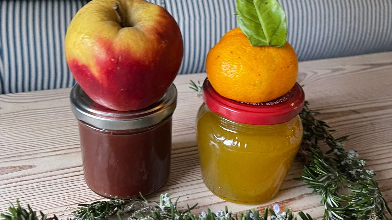 Two jars of jam with an apple and orange on top, placed on a wooden surface.