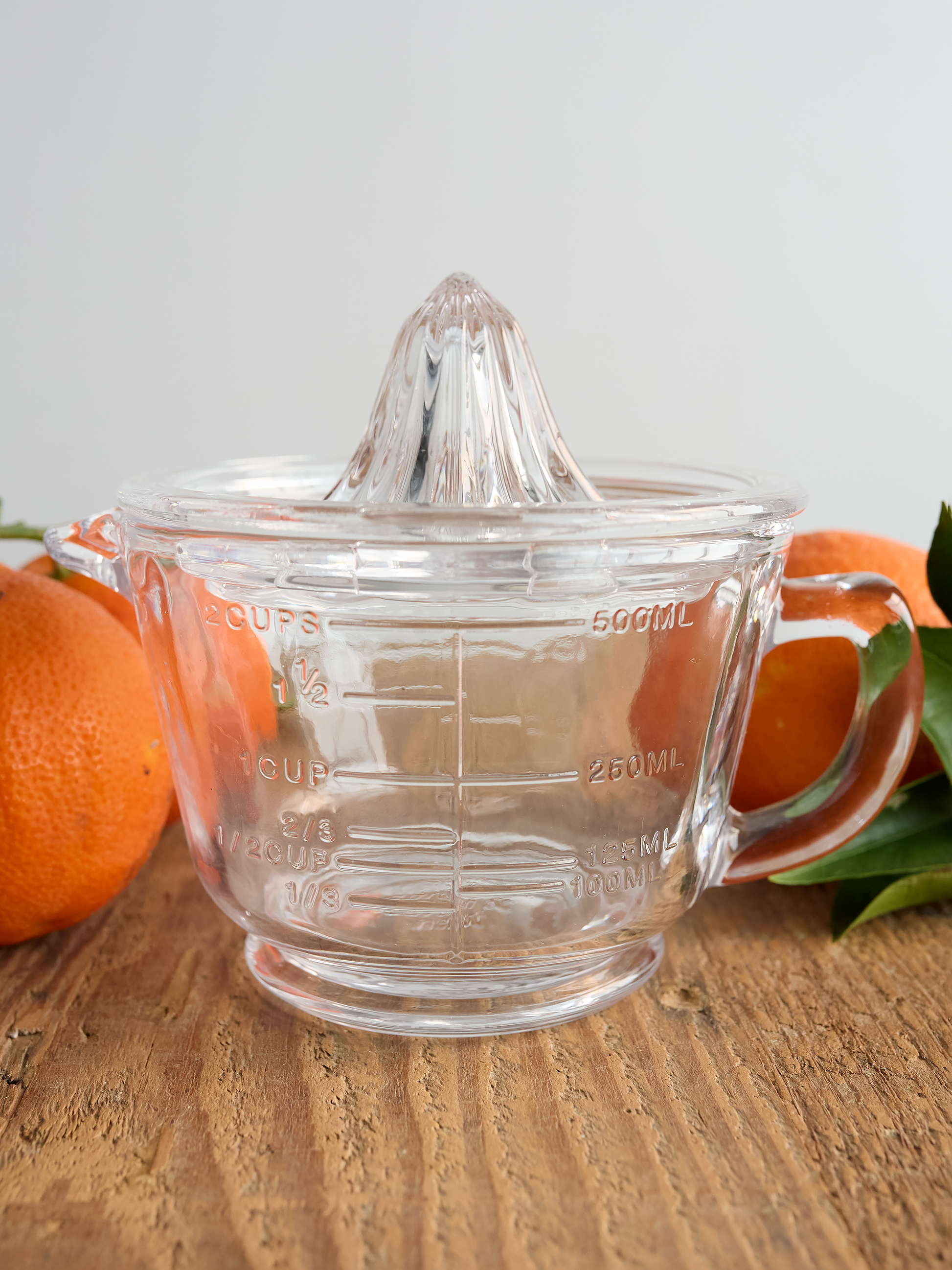 Clear glass juicer with measurement markings on a wooden surface with oranges in the background.
