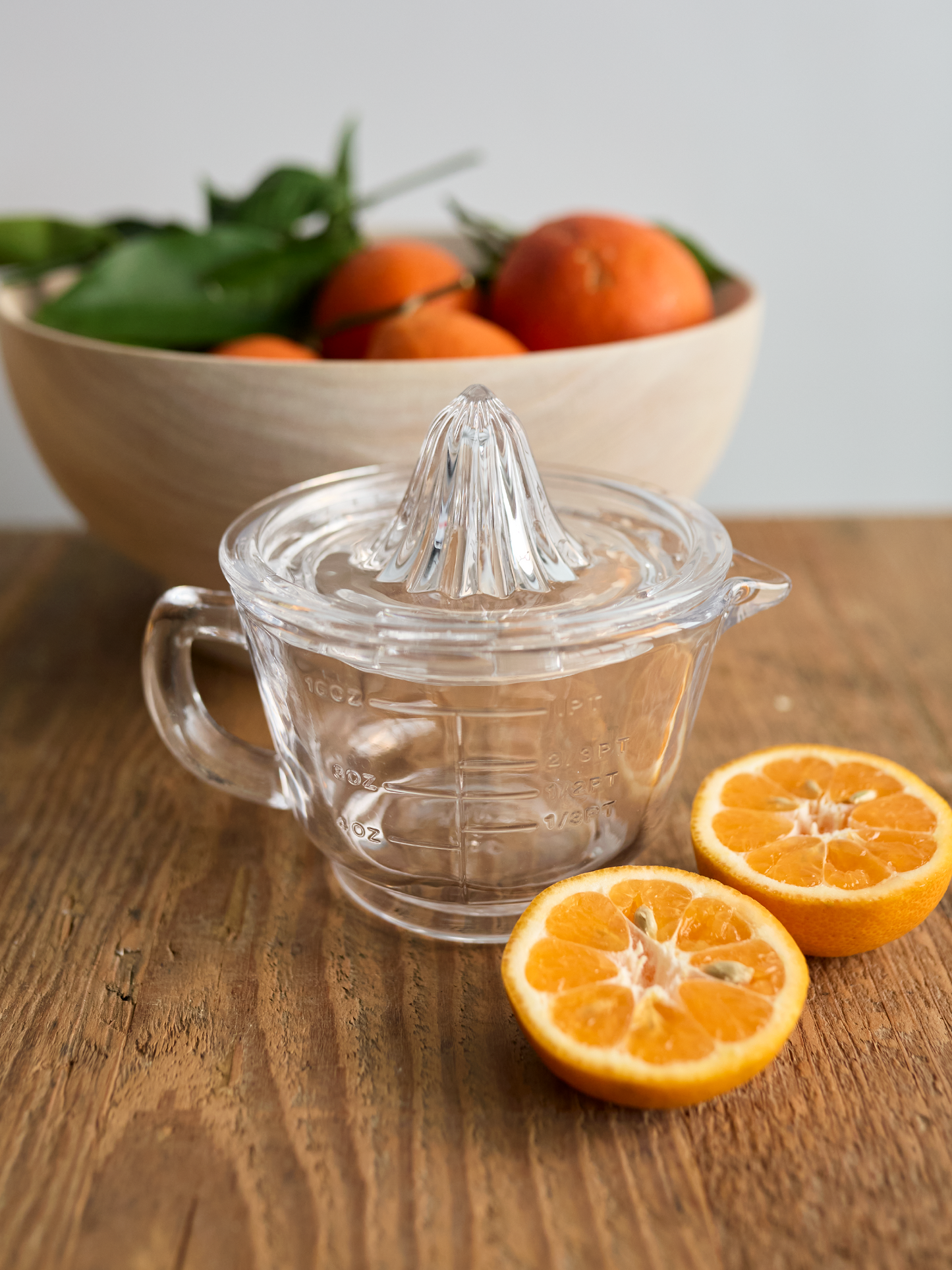 Clear glass manual juicer on a wooden table with oranges and a bowl of leaves in the background.