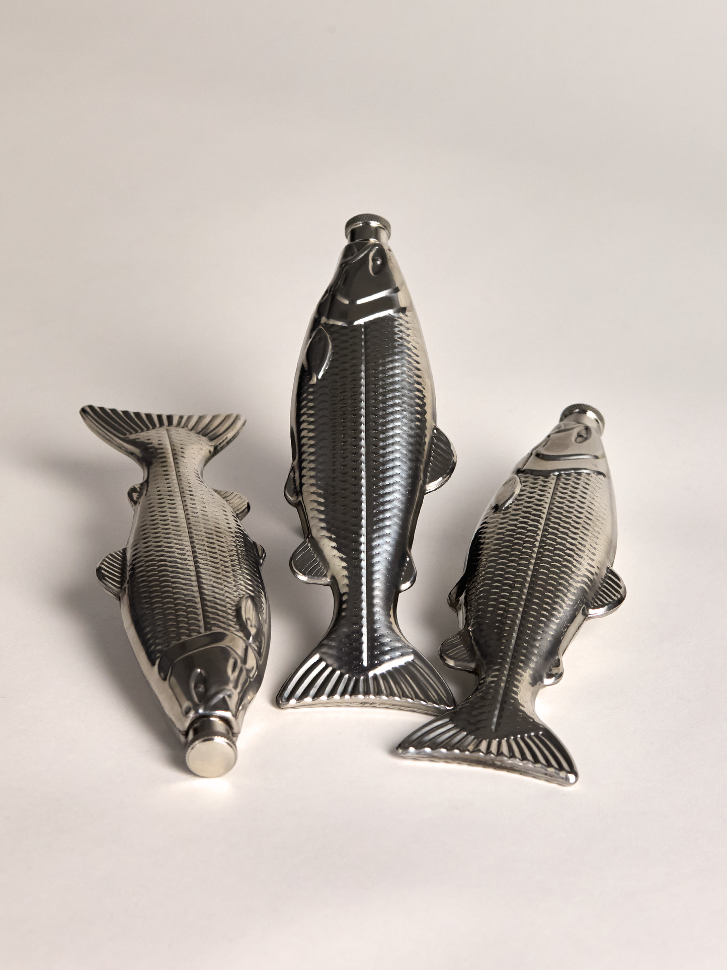 Three metallic fish-shaped flasks on a light gray background