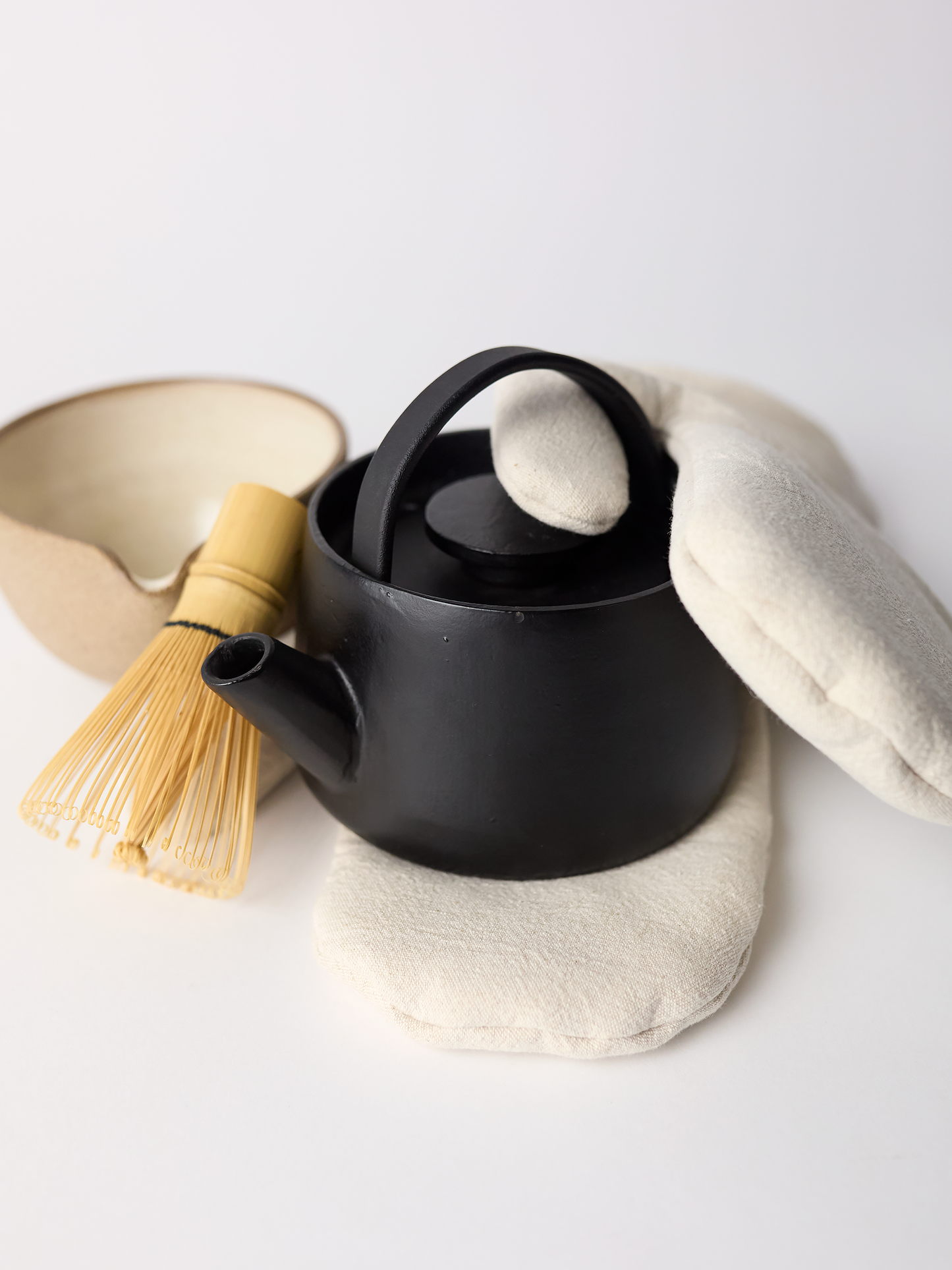 Black teapot with a wooden whisk ceramic bowl and mitts on a white background